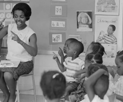 An adult is sitting in a chair at the front of a classroom and pointing in the air. Young children are seated on the floor around the adult and some are also pointing.