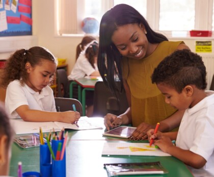 A teacher leans over a table in a brightly lit classroom, helping a young boy who is coloring with markers. Other children sit nearby working on their own drawings. The classroom is colorful and decorated with educational posters.
