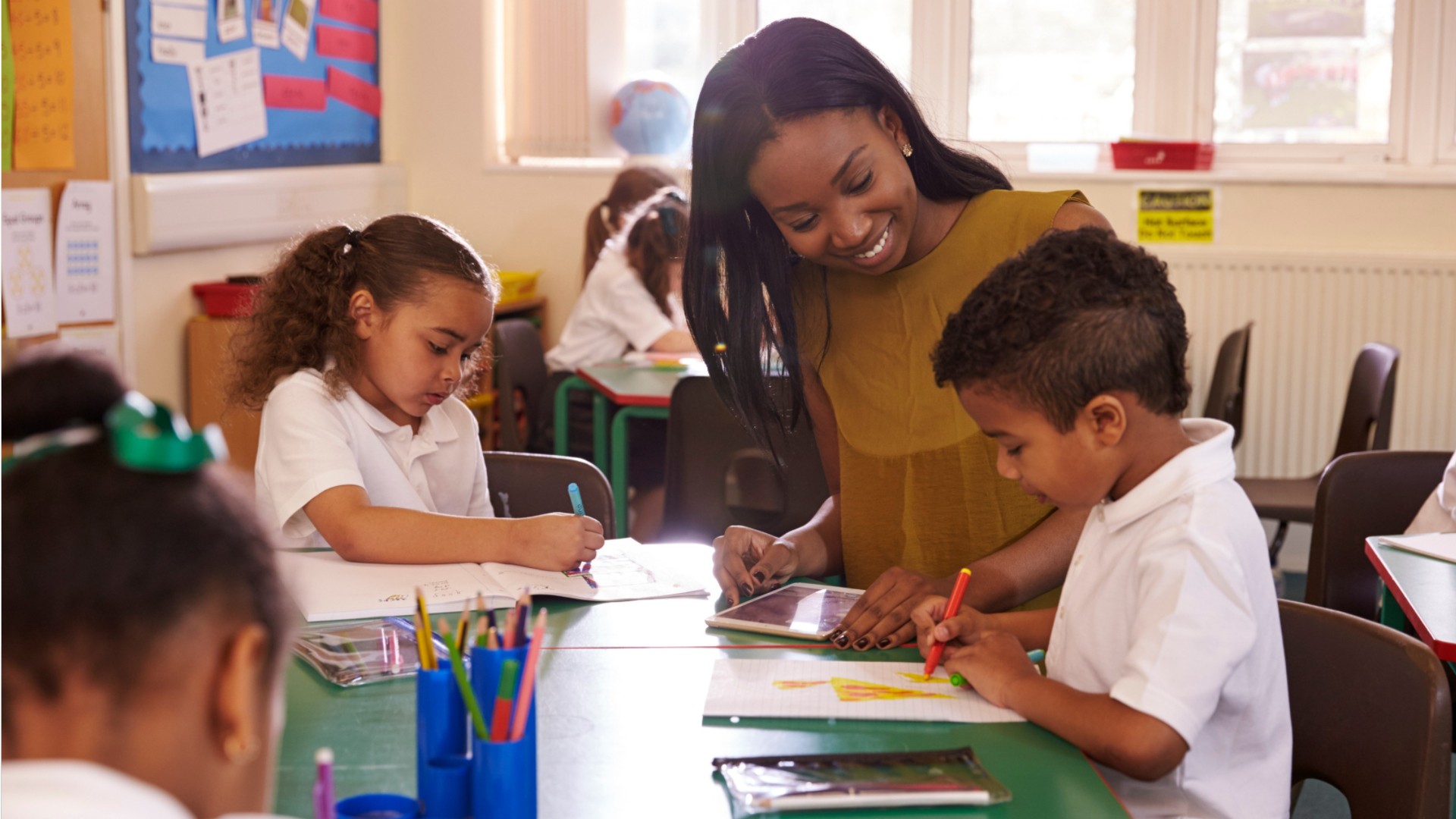 A teacher leans over a table in a brightly lit classroom, helping a young boy who is coloring with markers. Other children sit nearby working on their own drawings. The classroom is colorful and decorated with educational posters.